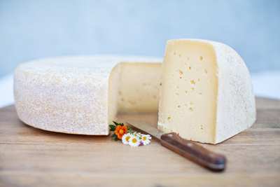 A large wheel of cheese with a wedge cut out, revealing a pale interior and a textured rind, placed on a wooden surface with a knife and small flowers.